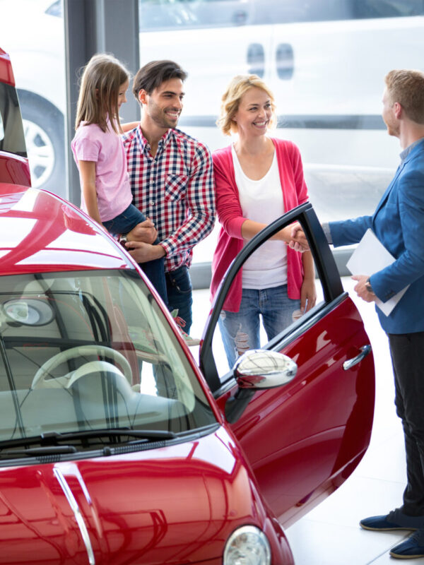 Smiling car agent showing vehicle to young family, all stand together next to the car in the car dealership  saloon